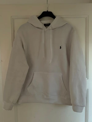 Vit hoodie - Snygg vit hoodie från Polo Ralph Lauren med klassisk huva och dragsnören. Har en stor magficka och den ikoniska lilla broderade loggan på bröstet. Perfekt för en chill och stilren look. Mjuk bomullskänsla och relaxed fit.