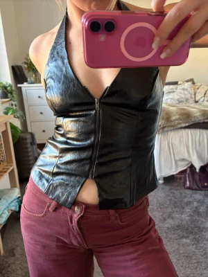 Svart halterneck topp i skinnimitation - Snygg svart halterneck topp i skinnimitation med dragkedja framtill och v-ringad urringning. Toppen är ärmlös och har en tight passform som framhäver figuren. Perfekt för dig som vill sticka ut med en edgy look.