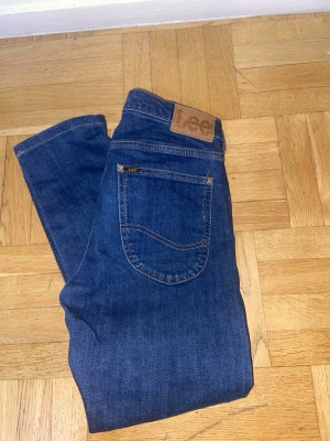 Blå jeans från Lee - Snygga blå jeans från Lee med klassisk femficksdesign och brunt läderpatch med logga bak i midjan. Jeansen har raka ben och är gjorda i slitstarkt denimtyg. Perfekta för en avslappnad och trendig look.