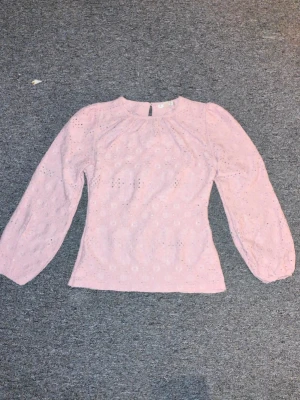 Ljusrosa/lila blus med detaljer - Supersöt ljusrosa blus med hålmönster och rund halsringning. Blusen har långa puffärmar och en liten knapp i nacken. Perfekt för en romantisk och feminin look.
