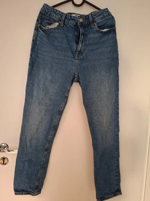 Blå mom jeans från H&M - Säljer ett par klassiska blå mom jeans från H&M i storlek 38. Jeansen har hög midja, raka ben och femficksmodell. Tillverkade i denim med snygg tvätt och knappgylf. Perfekta för en avslappnad och trendig look.