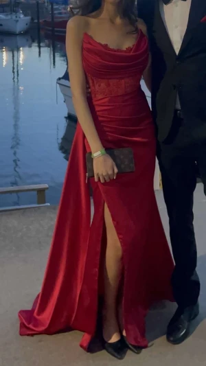 Röd lång balklänning med slits och spets - Elegant röd långklänning i glansigt tyg med figurnära passform, slits framtill och draperad överdel. Vackra spetsdetaljer och klänningen är axelbandslös med bred urringning. Använd endast en gång på bal. Storlek 34 Jag är 177 cm lång och bar klackar på bilderna klänningen är fortfarande lång, så passar även dig som är lång eller vill ha klackar. Tyget har lätt stretch och formar sig fint efter kroppen. Köpt på Proms & Weddings för 3500 kr, säljes för 2900, priset kan diskuteras.