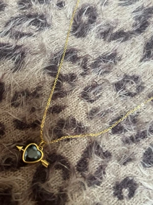 Guldigt halsband med hjärthänge svart hjärta - Snyggt halsband med tunn guldfärgad kedja och ett hjärtfomat hänge i svart sten, inramat i guld. Hänget har en pil genom sig vilket ger en cool och lekfull vibe. Perfekt accessoar för att lyfta din outfit med lite extra edge. Rostfri 