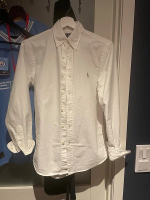 Vit skjorta från Ralph Lauren - Klassisk vit skjorta från Ralph Lauren med button-down krage och broderad logga på bröstet. Skjortan har långa ärmar och stängs med vita knappar framtill. Perfekt för en clean och stilren look.
