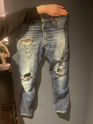 Blå slitna jeans med hål - Säljer ett par blå jeans med flera slitningar och hål på benen. Jeansen har en klassisk femficksmodell och smal passform. Perfekta för en avslappnad och trendig streetstil.