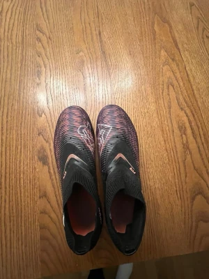 Puma Future svarta fotbollsskor - Säljer ett par svarta Puma Future fotbollsskor med röda detaljer och mönstrad ovandel. Skorna har strumpliknande skaft och röda dobbar för bra grepp på planen. Perfekta för dig som vill ha stil och funktion på fotbollsplanen.