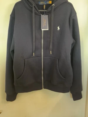  Polo Ralph Lauren zip  - Helt ny hoodie från Polo Ralph Lauren med dragkedja, huva med dragsko och klassisk logga broderad på bröstet. Mjuk bomullsblandning och två stora fickor framtill. Perfekt för en chill och stilren look.