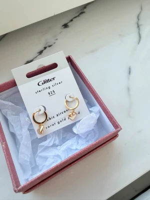 Guldpläterade örhängen med hjärta Glitter - Snygga örhängen från Glitter i 925 sterling silver med 18 karats guldplätering. Örhängena har en rund creolform och ett hängande hjärta i blank guldfinish. Perfekta för dig som gillar trendiga och söta accessoarer.