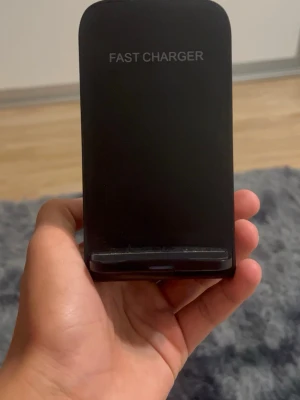 Svart trådlös snabbladdare - Stilren svart trådlös snabbladdare med texten 'FAST CHARGER' på framsidan. Laddaren har en upprätt design och är tillverkad i plast. Passar de flesta mobiler som stödjer trådlös laddning. Enkel att använda på skrivbordet eller nattduksbordet.