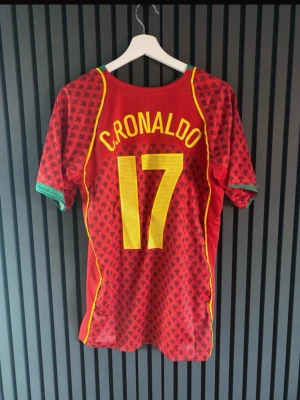Portugal fotbollströja C. Ronaldo 17 - Röd Portugal fotbollströja från Nike med C. Ronaldo och nummer 17 på ryggen. Tröjan har gula detaljer, gröna muddar och ett mönster över hela tyget. Landslagets emblem på bröstet och Nike-logga. Perfekt för fotbollsfans!