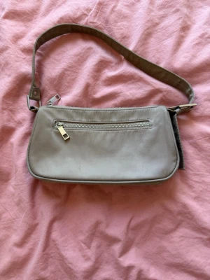 Beige axelremsväska med dragkedja från BikBok - Stilren beige handväska i avlång modell med justerbar axelrem och en praktisk dragkedjeficka framtill. Väskan har silverfärgade metalldetaljer och är tillverkad i ett slätt material som ger en clean look.