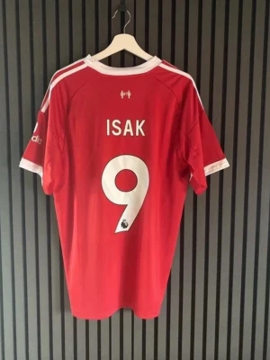 Liverpool matchtröja röd Adidas - Röd Liverpool FC matchtröja från Adidas med vita detaljer och tryck. Nummer 9 och namnet ISAK på ryggen, samt Premier League-logga. Framsidan har klubbmärke, Adidas-logga och sponsortryck. Perfekt för fotbollsfans!
