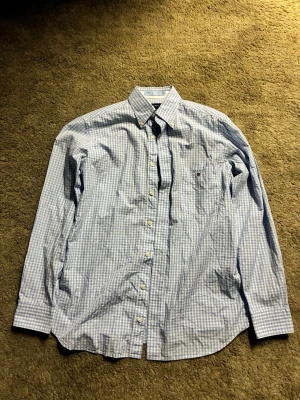 Blå skjorta från Gant - Klassisk blåvit rutig skjorta från Gant med lång ärm och knappar framtill. Skjortan har button-down krage och en liten logga broderad på bröstfickan. Perfekt för en stilren och avslappnad look.