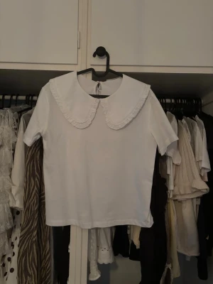 Vit blus med stor krage och volang - Supersöt vit blus med korta ärmar och en stor, rundad krage med volangkant. Blusen har en rak passform och knäppning bak i nacken. Endast använd 1 gång 💕