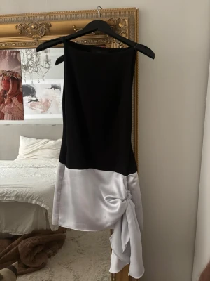 Svart och vit kort klänning med knyt - Säljer min älskade kort klänning i svart och vitt med unik knytdetalj på sidan. Klänningen har en rak, ärmlös design och är tillverkad i ett glansigt material som ger en lyxig känsla. Perfekt för dig som vill sticka ut med en modern look.