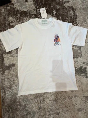 Vit Casa Blanca t-shirt med tryck - Vit t-shirt från Casa Blanca med färgglatt grafiskt tryck och logga på både bröstet och ryggen. Klassisk passform med rund hals och korta ärmar. Perfekt för dig som gillar streetwear och unika prints.