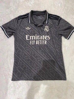 Grå Real Madrid Adidas tröja - Snygg grå fotbollströja från Adidas x Real Madrid med mönstrad design, klassisk krage och svarta ränder på axlarna. Tryck med 'Emirates Fly Better' på bröstet och klubbmärke. Perfekt för träning eller match.