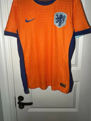 Holland fotbollströja orange Nike - Snygg orange fotbollströja från Nike med Nederländernas landslagsemblem på bröstet. Tröjan har mörkblå detaljer på sidorna och ärmarna samt rund halsringning. Tillverkad i lätt och ventilerande material, perfekt för träning eller match.
