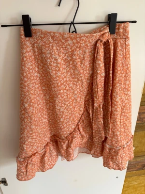 Orange omlottkjol med volang - Söt orange omlottkjol med små vita blommor och volangkant. Kjolen har knyt i sidan och är perfekt för en trendig och avslappnad look. Lätt och luftig modell som passar till många tillfällen.