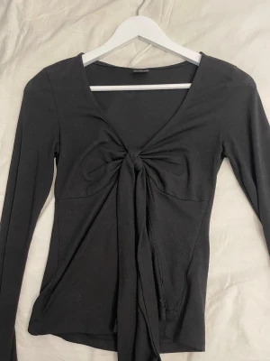 Svart långärmad topp med knyt Gina Tricot - Svart långärmad topp från Gina Tricot i storlek XS. Toppen har v-ringning och snygg knytning framtill som ger en trendig look. Perfekt att styla med jeans eller kjol för en cool outfit.