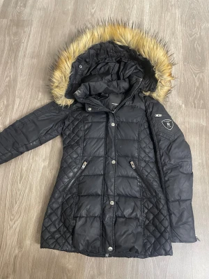 Svart Rockandblue dunjacka med päls - Svart dunjacka från Rockandblue med snygg quiltad design och fluffig päls på huvan. Jackan har dragkedjor på fickorna, tryckknappar framtill och en patch på ärmen. Perfekt för kalla dagar med sin långa modell och varma känsla.  jag sänkte priset från 3500kr till 900 och kan sänka mer PRISET GÅR ATT DISKUTERA!!