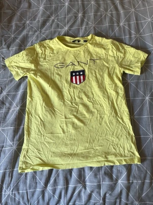 Gul t-shirt från GANT med tryck - Gul t-shirt från GANT med klassiskt tryck och logga på bröstet. T-shirten har rund halsringning och korta ärmar. Perfekt för en avslappnad stil och enkel att matcha med jeans eller shorts.
