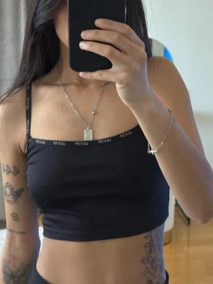 Levi's topp - Svart croppad linnetopp från Levi's med smala axelband och logotypdetaljer längs kanterna. Toppen har en tight passform och är tillverkad i ett mjukt, stretchigt material