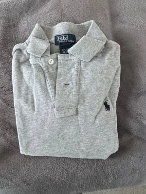 Grå pikétröja från Polo Ralph Lauren - S i barnstorlek! Klassisk grå pikétröja från Polo Ralph Lauren med broderad logga på bröstet. Tröjan har korta ärmar, krage och knäppning framtill. Tillverkad i mjuk bomull som känns skön mot huden. Perfekt för en stilren och avslappnad look.