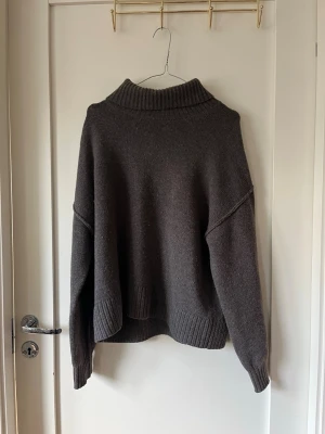 Mörkbrun stickad polotröja - Mysig mörkgrå / mörkbrun stickad polotröja med bred ribbad krage, ärmslut och nederkant. Tröjan har en avslappnad passform och långa ärmar, perfekt för kyliga dagar. Materialet ser ut att vara ull eller ullblandning.