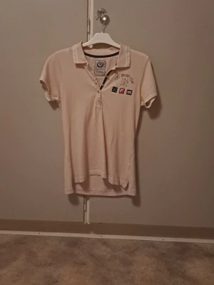 Ljusrosa pikétröja från Etirel - Ljusrosa pikétröja från Etirel med broderad text 'NYC Yacht Club' och flaggor på bröstet. Klassisk krage med knappar och diskreta ränder på kragen. Perfekt för en sportig och avslappnad stil. Ger English sophisticated schoolgirl vibe, 2000s horsegirl vibe, scandi style vibe o Stockholm vibe