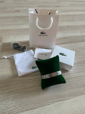  Lacoste armband - Snyggt armband i silverfärgad metall från Lacoste med rektangulära länkar och diskret krokodillogga. Armbandet har en stilren och modern design med mönstrade detaljer på länkarna. Perfekt accessoar för att lyfta din outfit.