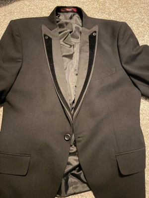 Svart kavaj med sammetsdetaljer - Stilren svart kavaj  med smala slag och snygga sammetsdetaljer längs kragen. Kavajen har en knapp framtill, två fickor med lock och en bröstficka. Perfekt för en dressad look och tillverkad i ett slätt tyg med fodrad insida.
