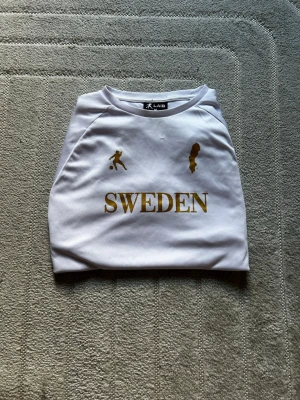 Vit Sverige t-shirt från Laib - Vit sportig t-shirt från Laib med guldiga detaljer, texten 'SWEDEN' på bröstet samt tryck av en fotbollsspelare och Sveriges karta. T-shirten har korta ärmar och gula samt blå ränder längs sidorna och ärmsluten. Perfekt för att heja fram Sverige!