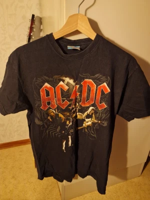 Svart AC/DC t-shirt från Starworld - Cool svart t-shirt med stort AC/DC-tryck på bröstet från Starworld. Klassisk rund hals och korta ärmar. Perfekt för dig som gillar rock och vill sticka ut med en ikonisk bandtröja. Materialet är mjuk bomull som känns skönt mot huden.