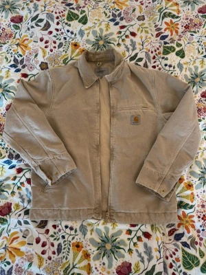 Beige Carhartt jacka med dragkedja - Säljer en beige Carhartt jacka i bomull med klassisk krage och dragkedja framtill. Jackan har en bröstficka med Carhartt-logga och guldiga knappar vid ärmsluten. Perfekt för dig som gillar streetwear och enkel stil.
