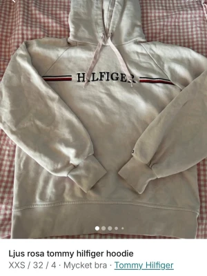 Ljusrosa hoodie från Tommy Hilfiger - Snygg ljusrosa hoodie från Tommy Hilfiger med broderad logga och ränder i svart, vitt och rött över bröstet. Klassisk huva med dragsko och ribbade muddar vid ärmslut och nederkant. Perfekt för en avslappnad och trendig look.