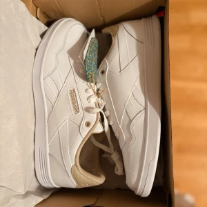 Vita Reebok Court Advance sneakers - Fräscha vita sneakers från Reebok, modell Court Advance. Skorna har en klassisk låg siluett, vita snören och diskreta detaljer i beige vid hälen. Tillverkade i syntetmaterial med stilrena sömmar och Reebok-logga på sidan.