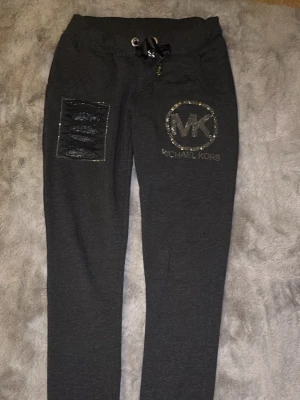 Gråa mjukisbyxor Michael Kors - Mörkgrå mjukisbyxor från Michael Kors med glittrande stenar runt MK-loggan på ena benet och en dekorativ ficka med svarta detaljer. Byxorna har dragsko i midjan och är i mjukt material, perfekta för en chill och snygg look.