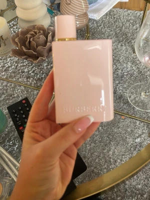 Burberry rosa parfymflaska, minimalt använd - Säljer en stilren parfym från Burberry i en ljusrosa, rektangulär flaska med gulddetalj vid halsen. Flaskan har en modern och minimalistisk design och är tillverkad i glas med en blank finish. Perfekt för dig som gillar eleganta dofter och snygg inredning på sminkbordet.
