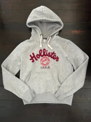 Hollister hoodie med tryck - Snygg trendig grå hoodie från Hollister med röd broderad logga och text på bröstet. Står storlek M i men skulle säga att de är en XS! Skriv vid funderingar💕