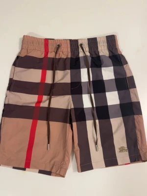 Rutiga shorts från Burberry - Snygga shorts från Burberry med klassiskt rutigt mönster i beige, svart, vitt och rött. Shortsen har snörning i midjan och en bakficka med knapp. Perfekta för sommaren och har en modern, loose passform.