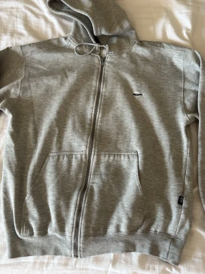 Grå zip hoodie från Oakley - Snygg grå zip hoodie från Oakley med dragsko i huvan och två stora fickor framtill. Tillverkad i mjukt bomullsmaterial med Oakley-logga på bröstet och en liten patch vid fickan. Perfekt för chill dagar eller när du vill ha en clean streetwear-look.