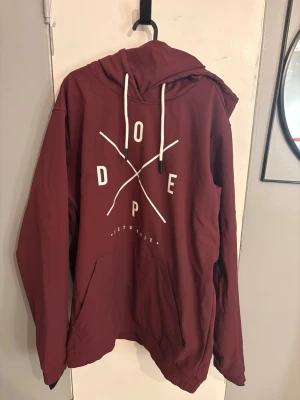 Vinröd jacka från Dope - Vinröd jacka från Dope med vit tryckt logga på bröstet och korsade linjer. Huvan har vit snörning och det finns en stor magficka. Ribbad nederkant och ärmslut ger en skön passform. 
