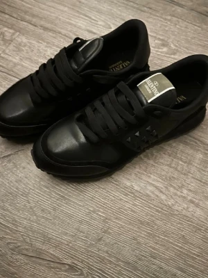 Svarta Valentino sneakers med nitar - Snygga helsvarta sneakers från Valentino med coola nitar på sidorna och breda svarta skosnören. Skorna har en klassisk låg modell och är gjorda i en mix av läder och textil. Perfekta för dig som vill ha en stilren men edgy look.