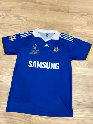 Chelsea Champions League tröja blå - Chelsea FC matchtröja i blått med vita detaljer från Adidas. Tröjan har Champions League-märke på ärmen, vit krage och Samsung som sponsortryck. Materialet är lätt och andas, perfekt för fotboll eller träning. Officiell klubbmärke på bröstet.