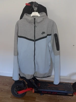 Nike grå hoodie med dragkedja - Nike hoodie i ljusgrått och grått med svart logga på bröstet. Hel dragkedja framtill, huva och svarta detaljer. Ficka med dragkedja på ena ärmen. Perfekt för en sportig och avslappnad stil.