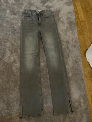 Grå raka jeans med slits - Säljer ett par grå raka jeans med hög midja och slitsar längst ner på benen. Jeansen har klassisk femficksmodell och är tillverkade i denim med en lätt tvättad look. Perfekta för en avslappnad och trendig stil.