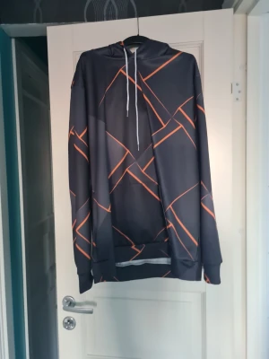 Svart hoodie med orange mönster - Snygg svart hoodie med grafiskt orange mönster och vita dragsnören i huvan. Hoodien har långa ärmar, ribbade muddar och en klassisk passform. Materialet ser ut att vara mjukt och lite glansigt, perfekt för en avslappnad streetwear-look.