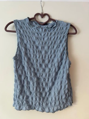 Blå ärmlös topp med struktur - Säljer en blå ärmlös topp med snygg struktur i tyget. Toppen har en rund halsringning och är tillverkad i ett lätt, våfflat material som ger en cool och avslappnad look. Perfekt att styla med jeans eller kjol för en trendig outfit.
