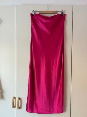 Axelbandslös långklänning i satin - Säljer en axelbandslös långklänning i starkt rosa satin. Klänningen har rak passform, slits baktill och en snygg knytning i ryggen. Materialet är glansigt och ger en lyxig känsla, perfekt för dig som vill sticka ut.